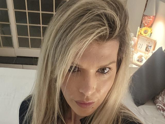 CherryBores has blonde, slightly messy hair covering one eye, brown eyes, and a small mole under her right eye. She's wearing a black top, looking straight at the camera.