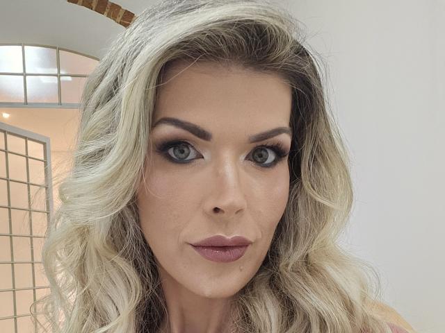 CherryBores tiene el cabello rubio y ondulado, ojos verdes y lápiz labial rosa. Su maquillaje incluye delineador de ojos oscuro y cejas bien definidas. Está mirando directamente a la cámara con una ligera sonrisa..