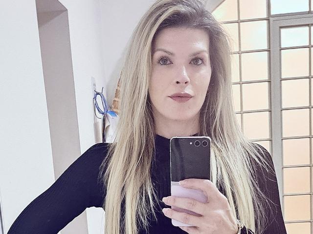 Eine Frau mit blonden Haaren, die ein schwarzes Oberteil mit langen Ärmeln trägt, hält ein schwarzes Telefon vor ihr Gesicht und macht ein Selfie im Spiegel. Ihre Brüste sind nicht sichtbar..