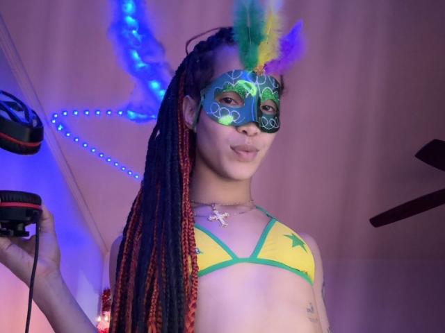 DolldSlov with long braids, wearing a colorful mask with feathers, yellow-green bikini, holding a camera, and a cross necklace, smiling with pouty lips, in a purple-lit room.