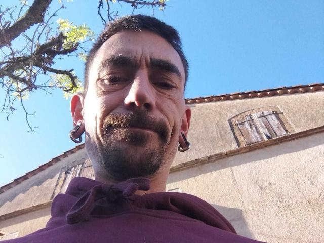 Xarly69, com cabelo curto e escuro, pequena barba e bigode, vestindo um moletom roxo, tem alargadores de orelha grandes e um leve sorriso, fotografado de baixo sob um céu azul claro..