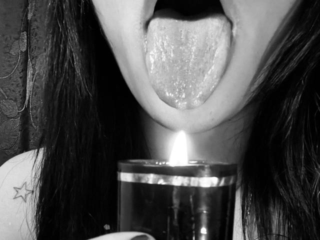 Close-up of FireCoupleHot's face with tongue out, sticking up towards a small lit candle. FireCoupleHot has dark, wet hair, and a visible star tattoo on their left shoulder. The candle's flame is centered in the image, casting light on FireCoupleHot's tongue and mouth. The image is black and white. The candle's glass is shiny and reflective. The texture of FireCoupleHot's tongue is moist and detailed. The overall composition is intimate and slightly provocative.