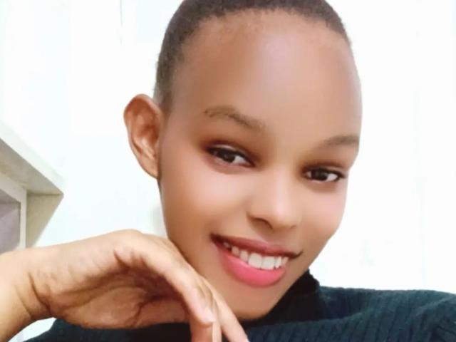 Le joli visage de Pascaliene, avec ses cheveux courts, sa peau brune et son rouge à lèvres rose, souriante et avec sa main sur son menton, portant un haut noir. Ses seins sont petits, et elle est attirante..