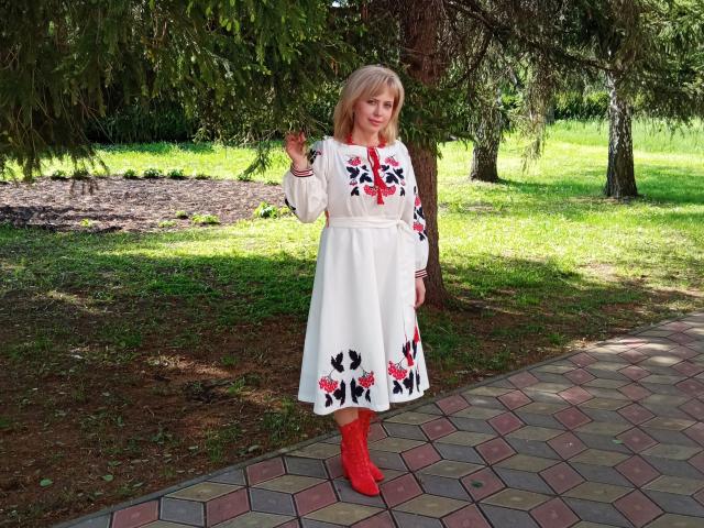 LaurenWishy stands in a sunlit park, wearing a white dress with long sleeves and red floral embroidery on the hem and chest. She has blonde hair and holds a small green leaf in her right hand. She's wearing red boots. The ground is a hexagonal pattern of bricks, and she's positioned near a tree with green foliage. The image has a bright, natural light.