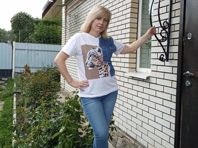 LaurenWishy stands in front of a brick house, right arm against the door. She has blonde hair, fair skin, and wears a white t-shirt with a blue denim pocket, featuring a leopard print graphic. She's also wearing blue jeans. Her left hand rests on her hip. The door has black wrought iron decor. Green plants and a fence are in the background. Her nails are painted red. The house has white brick with black lines. The overall style is casual.