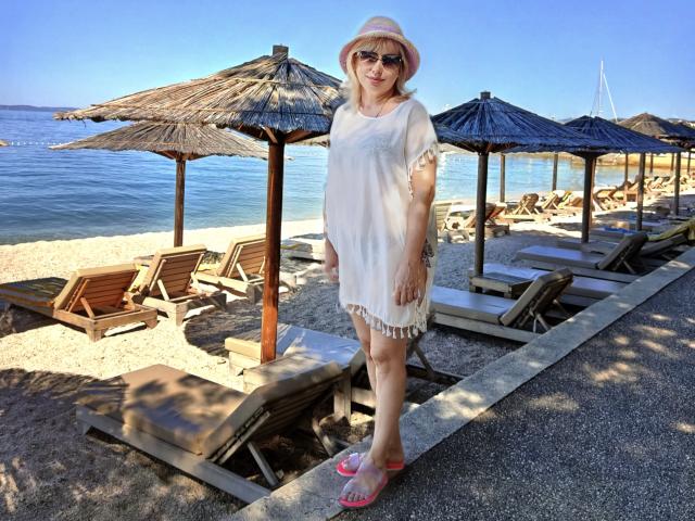 LaurenWishy stands on a gravel path in a beach setting. She wears a pink sun hat, dark sunglasses, a sheer white dress with frilled edges, and pink flip-flops. She's in front of several wooden beach chairs and thatched umbrellas. The background includes a clear blue sky, calm ocean, and sandy beach. The composition focuses on LaurenWishy, with the beach and ocean providing a serene backdrop. The texture of her dress is loose and airy. The image is brightly lit with natural sunlight.