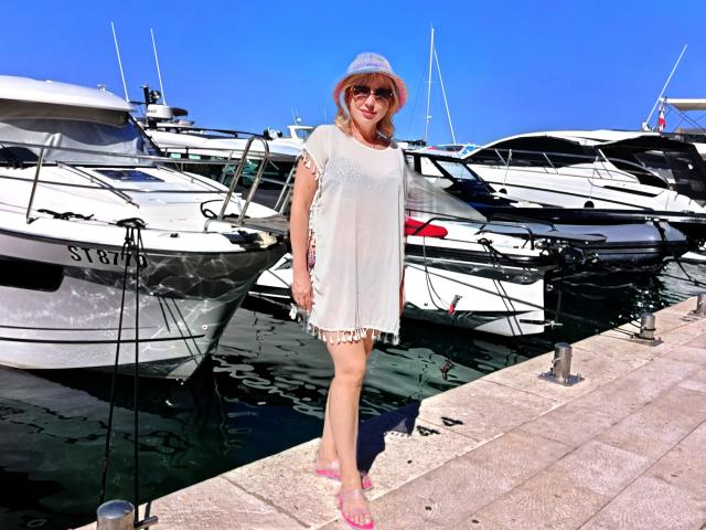 LaurenWishy stands in a marina, wearing a white, short, semi-sheer dress with frilled edges, and a light blue bucket hat. She has blonde hair, sunglasses, and pink sandals. Multiple white boats are docked behind her, with clear blue water and a bright, cloudless sky. The marina's stone-paved dock is in the foreground. LaurenWishy's relaxed pose and casual attire contrast with the sleek, polished boats. The image is vibrant and sunlit.