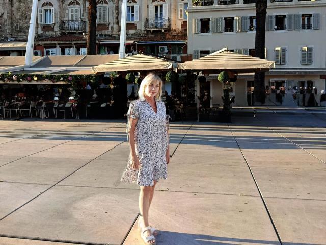 LaurenWishy stands in a sunlit square, wearing a short-sleeve, white floral dress with a V-neck. She has blonde hair, light skin, and visible tattoos on her right arm. She wears white sandals and stands with a relaxed, slightly forward posture. Two beige umbrellas are centered behind her, casting shadows on the square. The background includes a building with white shutters and a patio area with people seated. The square has a smooth, tiled surface. The overall composition is balanced with LaurenWishy in the foreground.