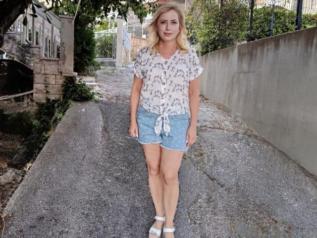 LaurenWishy stands on a cracked sidewalk, wearing a white floral blouse tied at the front, light blue denim shorts, and white sandals. She has blonde, shoulder-length hair and a slight smile. The image has natural light, and the background includes a stone wall, metal fence, and greenery. Her relaxed posture and casual attire contrast with the slightly worn sidewalk.