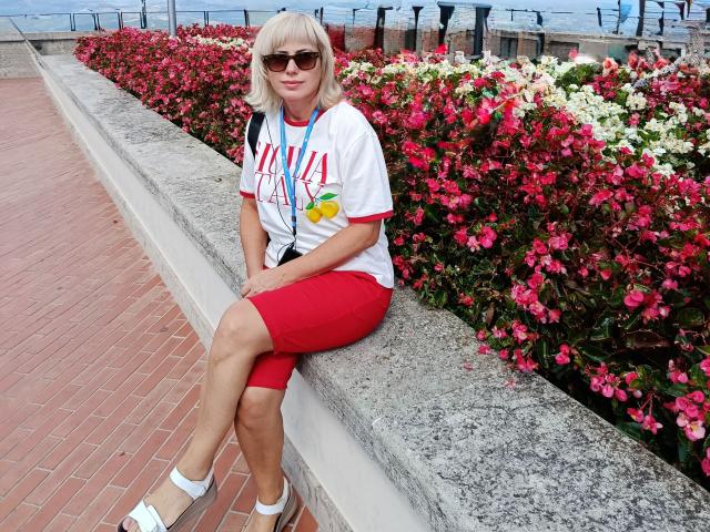 LaurenWishy sits on a stone ledge, wearing a white T-shirt with red lettering, red shorts, and white sandals. She has blonde hair, sunglasses, and a colorful lanyard. Bright pink and white flowers fill the background. LaurenWishy's legs are crossed, and she looks slightly to the right. The image has a watermark in the bottom right corner. The setting is a brick-paved waterfront.