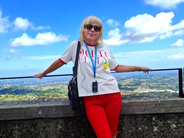 LaurenWishy stands against a stone railing with a clear view of a landscape. She wears a white T-shirt with "HOLLY" in red letters, red pants, and black sunglasses. A blue lanyard with a black ID card hangs from her neck. Her blonde hair is cut in a straight bang. She leans casually with one arm on the railing. Bright blue sky and fluffy white clouds are in the background. Her black shoulder bag is on her right side. The image is colorful and vibrant.