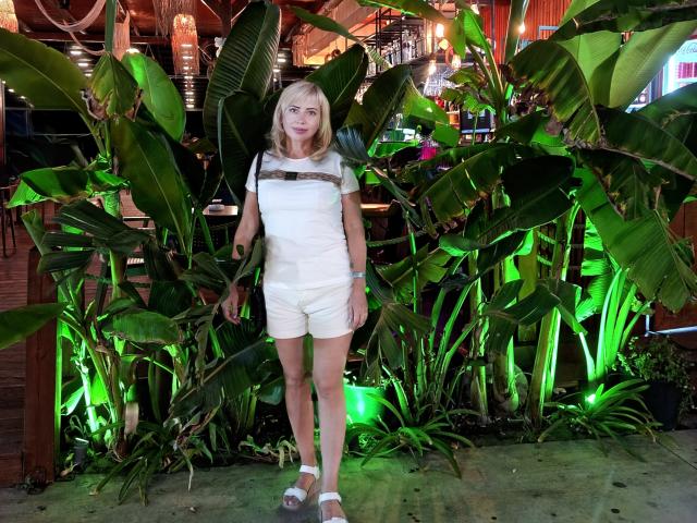 LaurenWishy stands in a tropical indoor setting, surrounded by large, green, leafy plants. She has blonde hair and a light skin tone, wearing a white short-sleeve top with a brown horizontal stripe and white shorts. She is also wearing white sandals. The background includes wooden elements and green lighting, with hanging lanterns and a bar area partially visible. The image has a natural, relaxed composition.