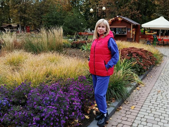 LaurenWishy stands on a brick path, wearing a bright red puffy vest, blue hoodie, and purple pants. She's in a garden with purple flowers, yellow grass, and red foliage. Her blonde hair is straight, and she's smiling. The background has a wooden hut and trees. The image uses natural colors and a casual, everyday setting. LaurenWishy's hands are in her pockets. The overall composition is relaxed and outdoor-friendly.