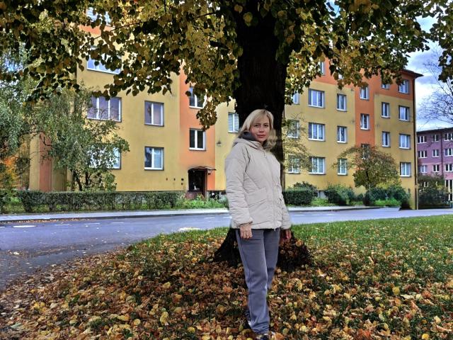 LaurenWishy stands in front of a large tree with yellow-green leaves. She wears a white puffy jacket and gray pants. The ground is covered in brown autumn leaves. The background shows a multi-story building with yellow and orange walls and white-framed windows. The sky is overcast. LaurenWishy has blonde hair and a slight smile. The image has a realistic style. The composition is balanced, with LaurenWishy centered and the tree dominating the upper part of the frame.