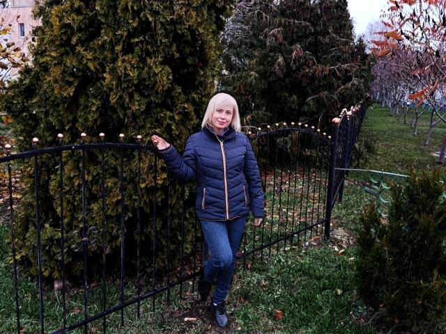 LaurenWishy stands in a yard, wearing a blue puffy jacket and blue jeans, with blonde hair, next to tall green bushes, waving at the camera.