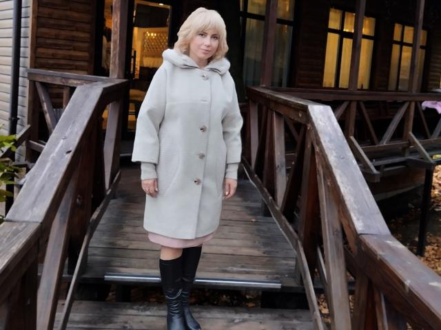 LaurenWishy stands on a wooden porch, wearing a light gray, button-up, woolen coat with a hood, black knee-high boots, and a pink skirt. She has short, wavy blonde hair and a slight smile. The wooden porch has a rustic design with dark brown railings and a small staircase. The background includes a wooden building with lit windows. The overall composition is simple, with a focus on LaurenWishy and the porch.