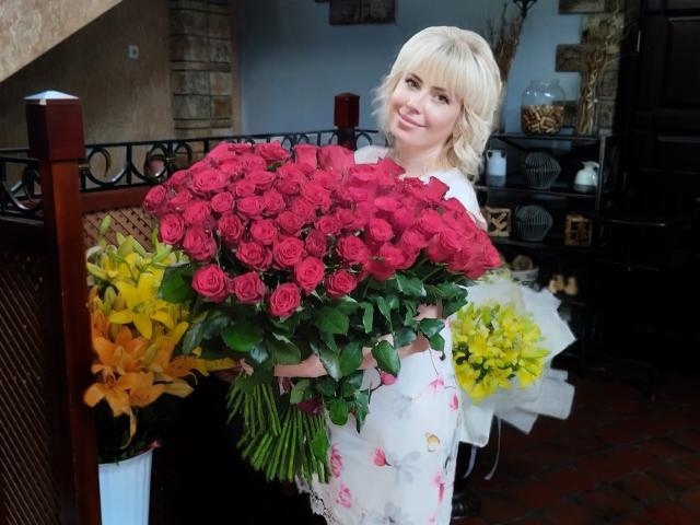 LaurenWishy with short blonde hair holds a large bouquet of red roses, mixed with yellow flowers and orange lilies. She wears a white floral dress, standing in a room with a black iron railing, brick floor, and decorative wall elements. Bright colors contrast with the room's darker background. Her smile is soft and relaxed. The bouquet is dense with roses, green leaves, and the yellow flowers adding variety. Image has a natural, candid feel.