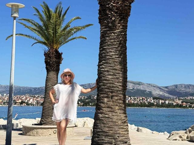 LaurenWishy stands next to a tall palm tree, wearing a white short-sleeve dress and a pink sun hat. She holds the tree with her right hand and has her left hand on her hip. Her skin is fair, and she wears sunglasses. The palm tree has a textured, rough bark. A streetlamp is to the left. In the background, there's a blue lake, a rocky shoreline, and a town with white and pink buildings. The sky is clear and blue. The image is bright and sunny.