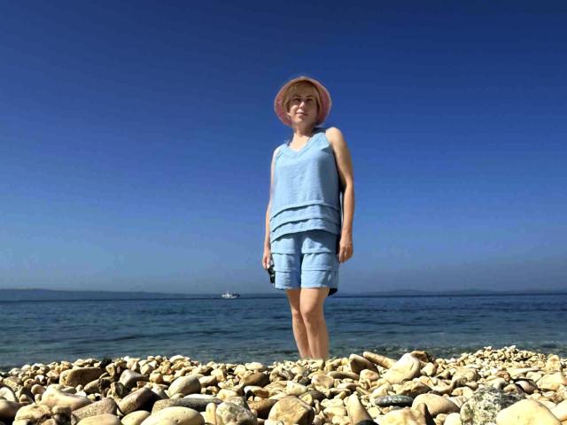 LaurenWishy stands on a rocky beach with clear blue water in the background. She wears a light blue sleeveless top and matching shorts, with a pink sunhat. Her skin is fair, and she has a relaxed expression. The rocks are varied in size and color, with smooth textures. A small white boat is visible on the water in the distance. The sky is bright blue, contrasting with the dark water. She stands slightly off-center to the right.
