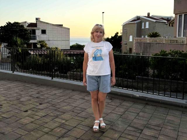 LaurenWishy stands on a brick-paved terrace, wearing a white T-shirt with a blue graphic, light blue denim shorts, and white wedge sandals. She has blonde hair and a slight smile. The background includes a black iron fence, green shrubs, and multi-story buildings at sunset. The sky is yellow-orange, and the ocean is visible in the distance. LaurenWishy is centered in the frame.