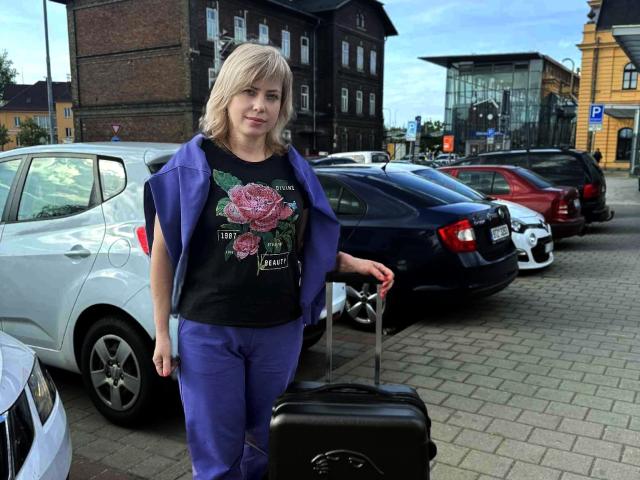 LaurenWishy stands in a parking lot, pulling a black suitcase with her right hand. She wears a black graphic t-shirt with pink roses and purple pants, with a blue jacket draped over her shoulders. She has blonde hair and a slight smile. Cars are parked in the background, including a white and a black car. The ground is paved with bricks. LaurenWishy is centered in the frame.