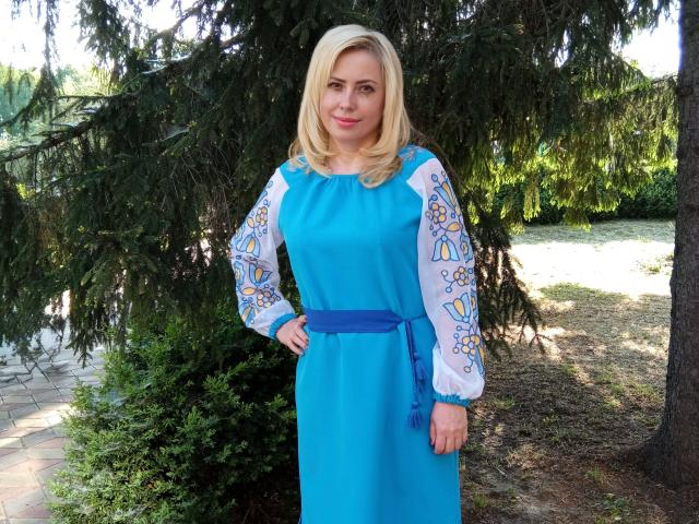 LaurenWishy stands outdoors, wearing a bright blue dress with white long sleeves featuring colorful floral patterns. Her blonde hair is shoulder-length, and she has a slight smile. She's positioned in front of a large, green, pine tree with dense foliage. The dress has a blue ribbon at the waist, and she's placed one hand on her hip. The background shows a grassy area and a stone pathway. The image is sharply focused, with natural light highlighting her features.