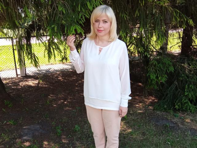 LaurenWishy stands in a wooded area, her blonde hair slightly wavy. She wears a white, long-sleeved blouse with a small, decorative knot at the chest and beige pants. She holds a green leaf with her right hand. Her expression is calm, with a slight smile. The sunlight filters through the trees, casting shadows on the ground. The image has a natural, casual feel. She is centered, with the foliage creating a dark green backdrop.