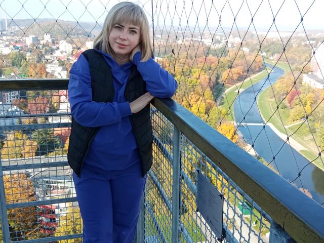 LaurenWishy stands on a metal balcony with a chain-link fence, leaning on the railing with her right hand. She has blonde hair, fair skin, and a slight smile. She wears a blue long-sleeve shirt, blue pants, and a black vest. The background shows a colorful autumn landscape with a river and buildings. The image is bright with clear, sharp details. LaurenWishy's casual pose and relaxed expression contrast with the structured fence and balcony.