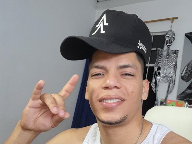 Andressant with a slight grin, wearing a black cap with a white "A" and white tank top, making a rock-and-roll hand gesture, showing a metal necklace.