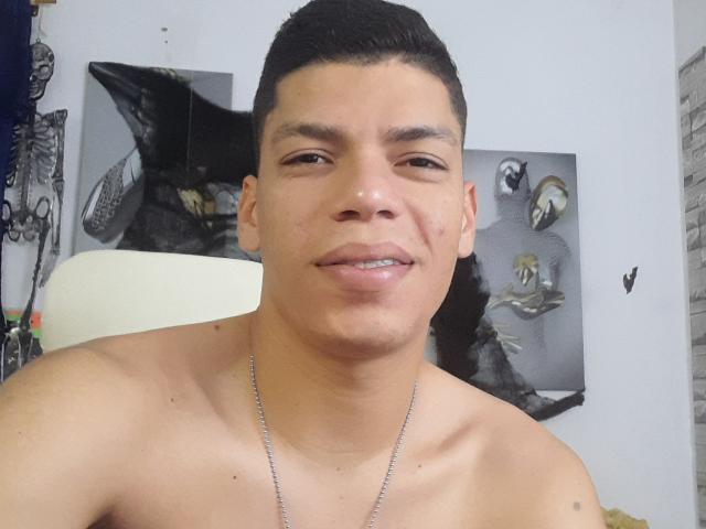 Andressant with short black hair, medium skin, and a slight smile, shirtless, wearing a silver necklace, has a small tattoo on his left shoulder. His ass is not visible.