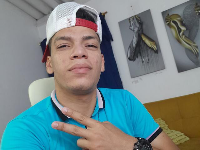 Andressant with light brown skin, wearing a white and red backwards cap, blue polo, and black watch, smirking, pointing at his neck with a finger.