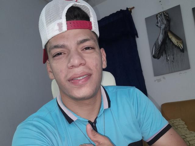 Andressant has a light brown skin tone, short black hair, and is wearing a white and red backwards cap with a blue polo shirt. He's smiling with his index finger up.