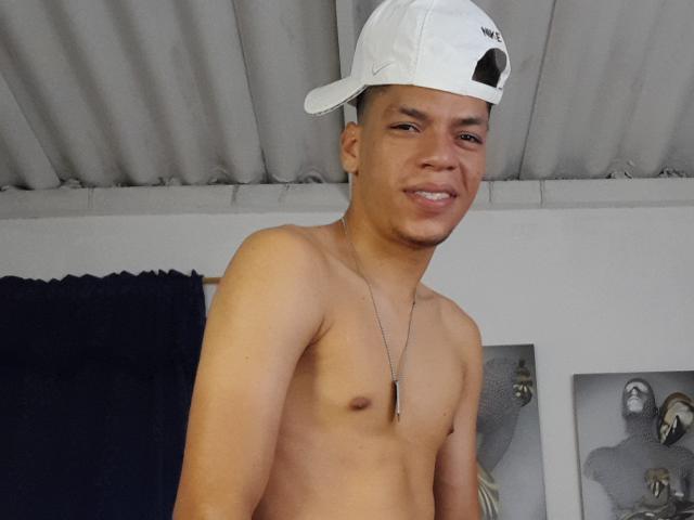 Andressant, shirtless with a small, cute ass, wearing a white cap and necklace, smiling slightly. He's a young, cute, dark-skinned dude with short hair.