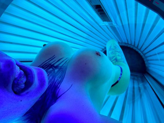 JuicyPeachy-hot lying in a tanning bed, blue lights, large tits, tattoo on chest, tongue out, purple skin, visible nipple, leg raised, arm partially visible, relaxed pose.