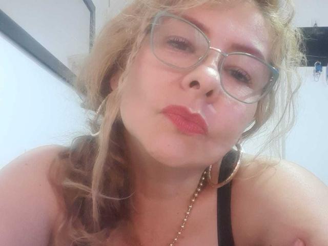 MiaAmoretti has pouty red lips, curly blonde hair, and wears glasses with hoop earrings, a black strap, and a gold necklace, making a cute face in a selfie.
