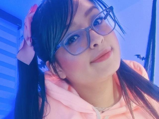 SofiiaThooner with glasses, pink hoodie, and pigtails, holding her hair up, smiling, blue-lit face, pink lips, and a cute, sexy look.
