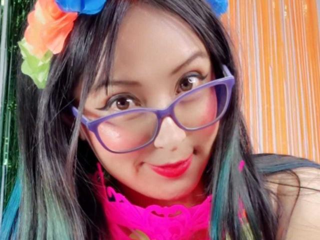 SofiiaThooner with purple glasses, black hair, blue and orange flower in hair, pink lace choker, and red lipstick, smiling at camera. Her tits are partially visible.