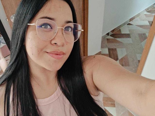 AudreyJoness with long black hair, wearing pink glasses and pink tank top, smiling, showing light skin and small acne on cheek, standing in doorway, holding out arm.