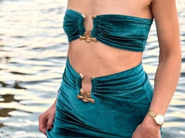 De groene fluwelen outfit van MiraGi met gouden kruisknopen, die haar borsten en billen laat zien, terwijl ze bij het water staat. Ze draagt een gouden horloge om haar rechterpols..