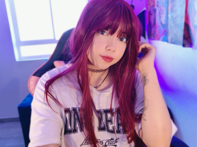 Arieyaara with red hair, blue eyes, wearing a choker and white "CONCERT" shirt, touching her hair, showing tattoos on her arm, sitting in a blue chair.