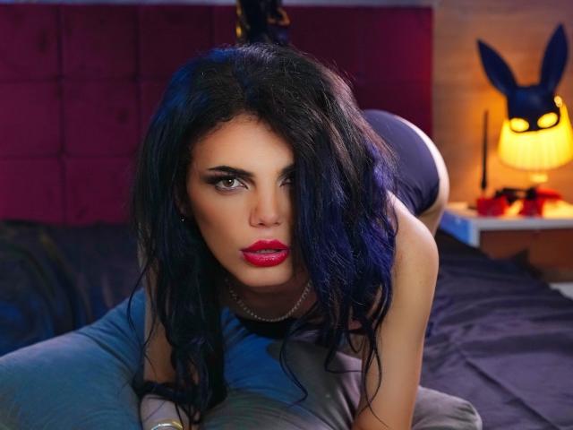 VanessaDomy is on all fours on a bed, looking directly at the camera with intense, dark eyes. She has long, wavy black hair and bright red lipstick. She wears a black sleeveless top and a pearl necklace. Her skin is fair. The background includes a dark purple headboard and a lit lamp with a Playboy logo. The bed has dark blue sheets. The image has a vibrant, slightly exaggerated color style. The composition is focused on VanessaDomy's face and upper body.