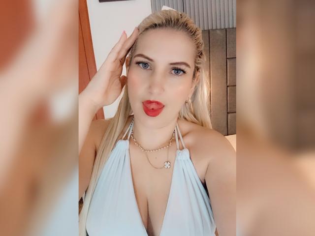 GiuliaAddams has blonde hair, blue eyes, and red lipstick. She's wearing a light blue halter top, showing her tits, and a pearl necklace. She's touching her head.