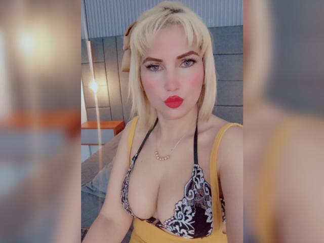 GiuliaAddams with blonde hair, red lipstick, and a black lace bralette showing her big boobs, wearing a yellow dress, and a gold necklace, looking at the camera.