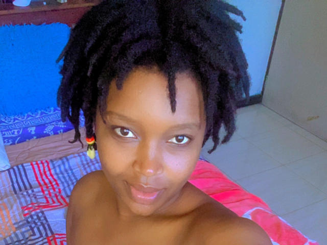 Close-up photo of SexyArya. She's a dark-skinned woman with short, curly black hair. Her eyes are wide and brown, and she's looking directly at the camera with a slight smile. She's topless, with visible shoulders. The background includes a blue wall, a wooden shelf, and a colorful bedspread with red, gray, and white patterns. She has small, dark nipples. The image has a natural, slightly blurry texture. Her ear has a colorful beaded earring.