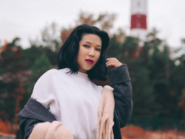 ErikaKim mit schulterlangem, schwarzem Haar, trägt ein weißes T-Shirt und eine graue karierte Jacke, einen pinken Schal, rote Lippenstift, die Hand ist in der Nähe des Gesichts, im Freien, bewölkt, weiches Licht, ein leichtes Lächeln..