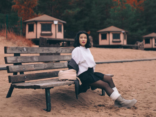 ErikaKim with short black hair, sitting on a wooden bench, wearing a white shirt, black skirt, black tights, white socks, and beige boots, with a pink bag on her left.