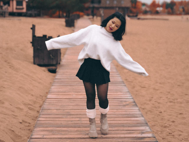 ErikaKim with black bob, white sweater, black skirt, black tights, white leg warmers, gray boots, dancing on wooden boardwalk, sandy beach, smiling, arms out, flirty pose.