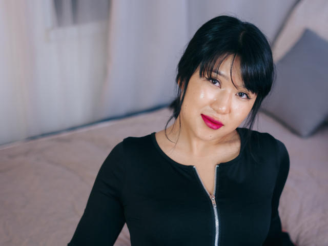 ErikaKim has black hair, red lipstick, and a shiny face, wearing a black zip-up top, with medium tits visible, sitting on a bed with a pillow in the background.
