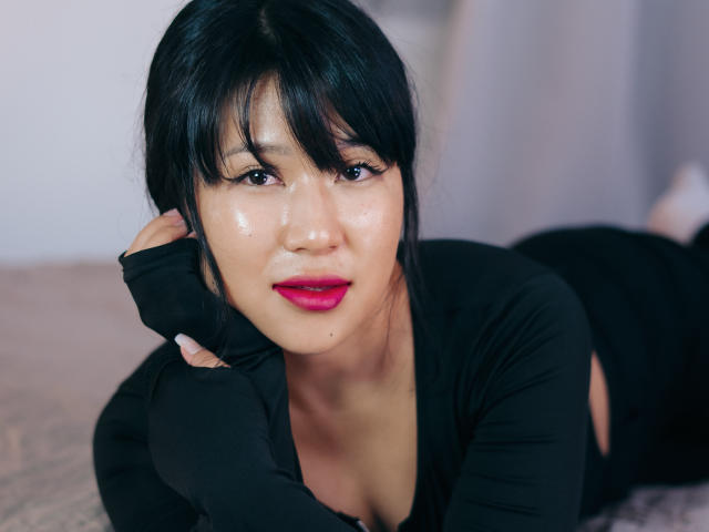 ErikaKim, avec une peau éclatante, des cheveux noirs, du rouge à lèvres et une tenue noire, allongée sur un lit, son visage brillant, une main touchant sa joue, regardant la caméra, ses seins partiellement visibles..