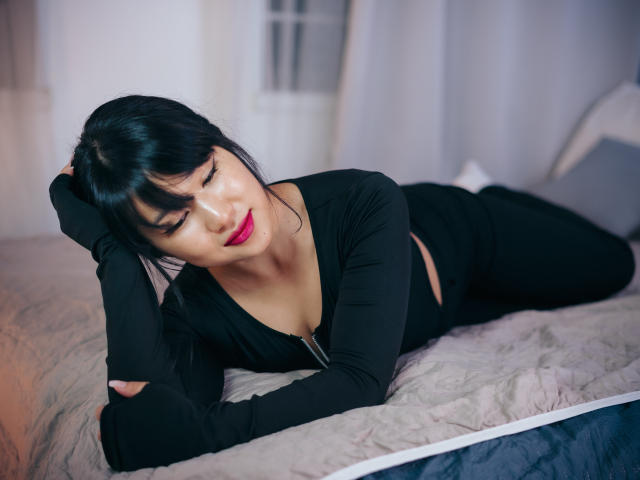 ErikaKim, liggend op bed, zwarte top met lange mouwen, zwarte broek, decolleté zichtbaar, roze lippenstift, zwart haar, pony, rechterarm onder haar hoofd, linkerarm rustend op het bed, lachend..